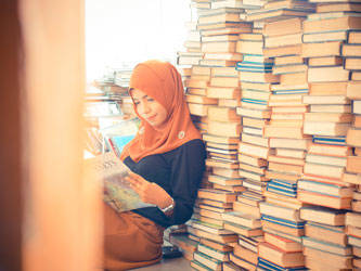 A Woman reading a book, sitting by a large stack of books